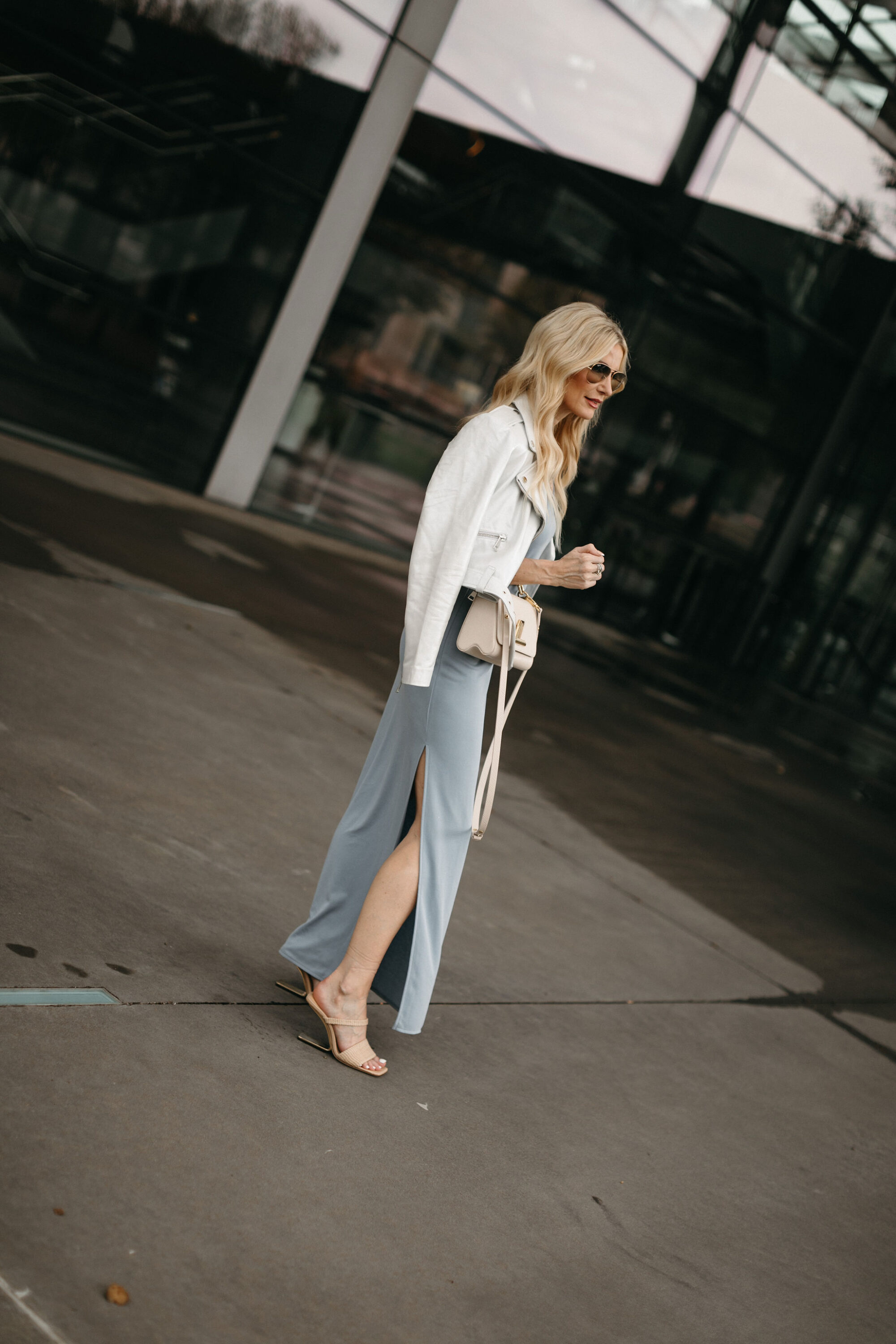Over 40 woman from Dallas wearing a white leather jacket over top a light blue knit midi dress with neutral heeled sandals.